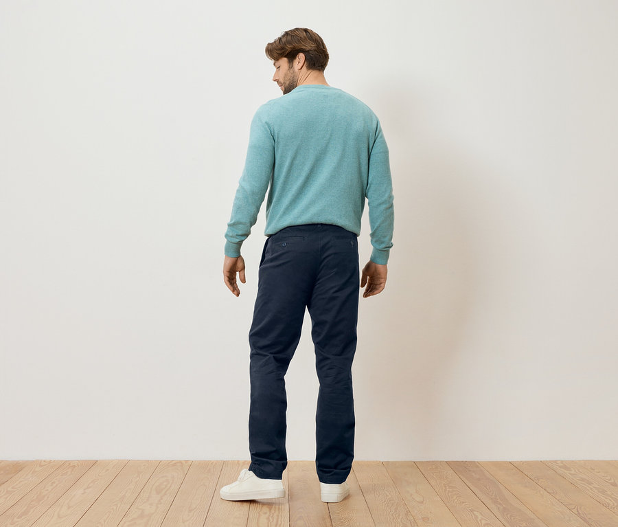 Ein Mann steht mit dem Rücken zur Kamera, er trägt einen mintfarbenen Pullover, dunkelblaue Hosen und weiße Turnschuhe.