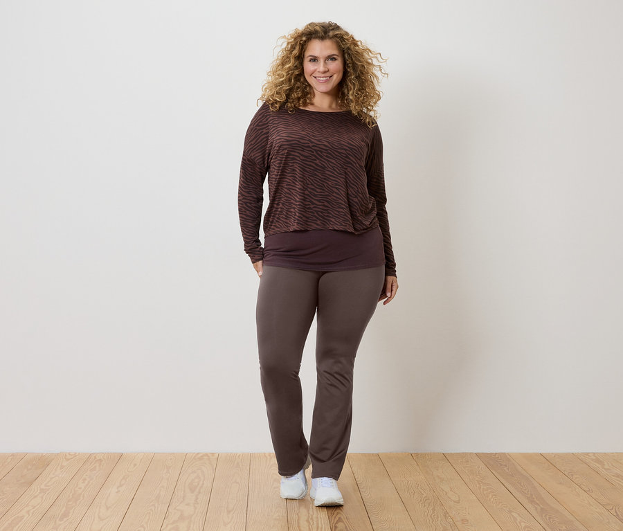Frau mit lockigem Haar trägt ein braun gemustertes Langarmshirt, ein dunkleres Top und braune Hosen, dazu weiße Turnschuhe.