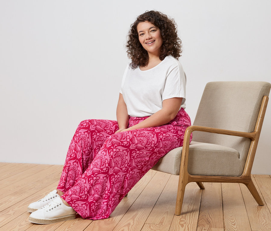 Frau mit lockigem Haar sitzt in einem beigen Sessel und trägt ein weißes T-Shirt, eine rosafarbene Hose mit Paisleymuster und weiße Turnschuhe.