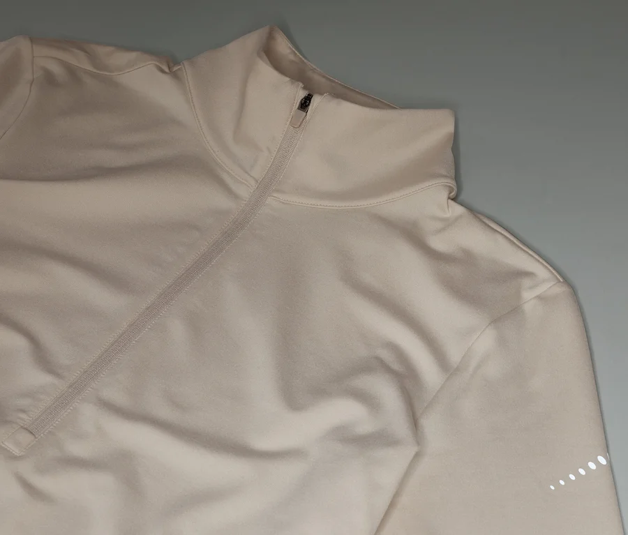 Beige Sportjacke mit Reißverschluss und hohem Kragen liegt auf grauem Hintergrund.