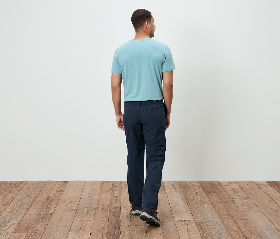 Mann von hinten, trägt ein türkisfarbenes T-Shirt und marineblaue Hosen, der auf einem Holzboden geht.