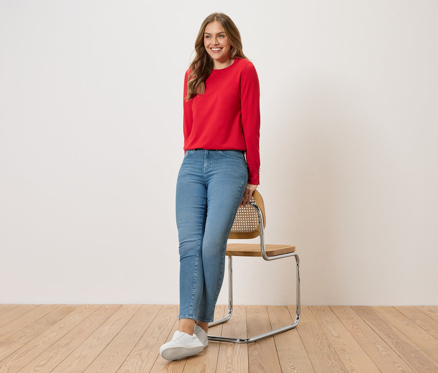 Eine Frau lehnt an einem Stuhl. Sie trägt einen roten Pullover, blaue Jeans und weiße Turnschuhe.