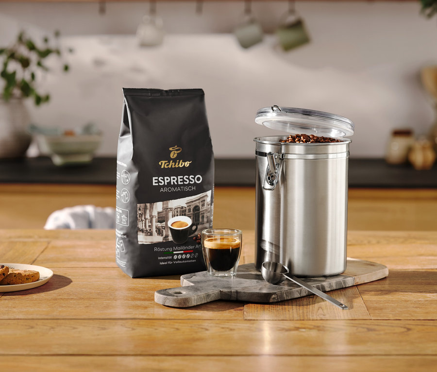 Espressopackung steht neben geöffneter Aromadose aus Edelstahl und Tasse auf einem Holztisch.