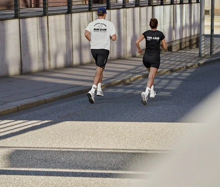Ein Mann in weißem Sportshirt mit „RUN CLUB“-Aufdruck, schwarzen Sportshorts und Unisex Coffee Crew Tennissocken und eine Frau in schwarzem Sportshirt mit „RUN CLUB“-Aufdruck und kurzen ActivePower Sportleggings laufen auf einer Straße.