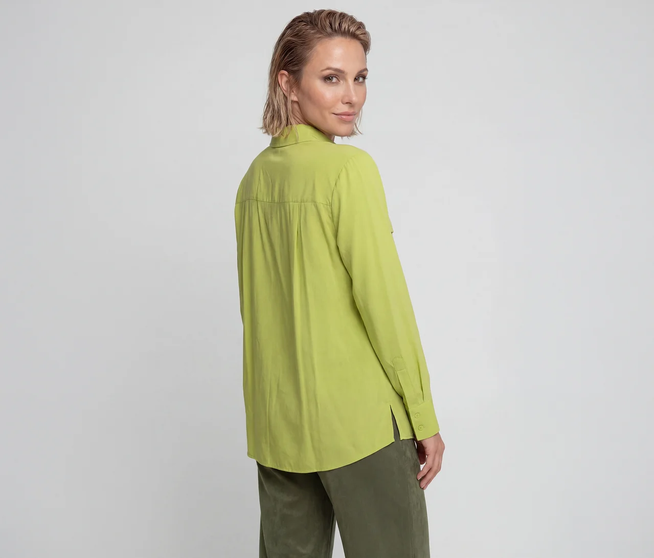 Frau mit kurzen blonden Haaren, von hinten gesehen, blickt über ihre Schulter. Sie trägt ein limettengrünes Langarmshirt und olivgrüne Hose.