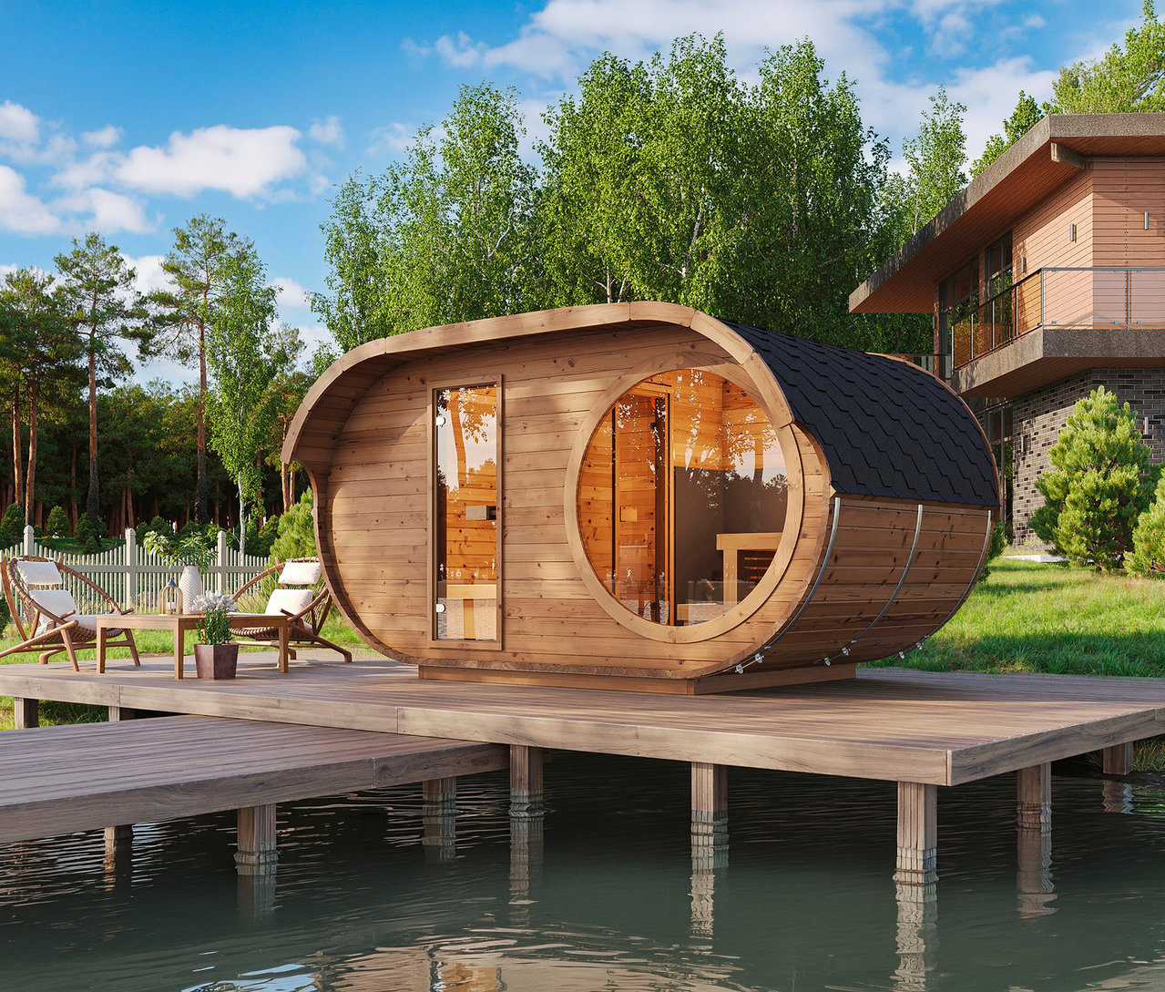 Holzfasssauna mit Glastür und rundem Fenster auf Holzterrasse über Wasser, daneben Gartenmöbel, umgeben von Bäumen und Haus.