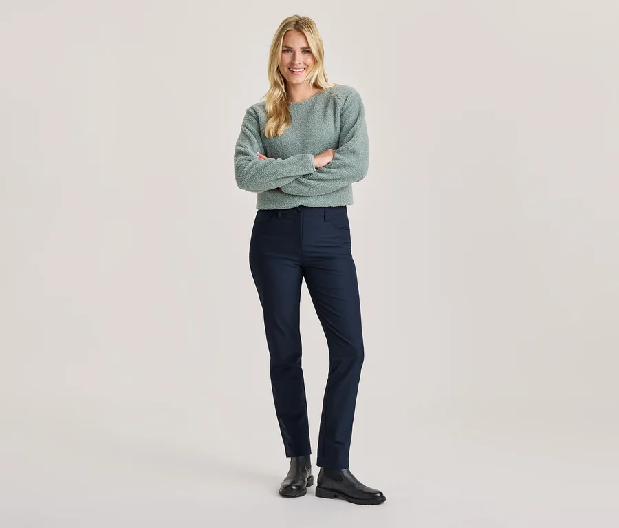 Lächelnde Frau mit blonden Haaren, verschränkte Arme, in hellgrünem flauschigem Pullover, dunkelblauer Hose und schwarzen Stiefeln.