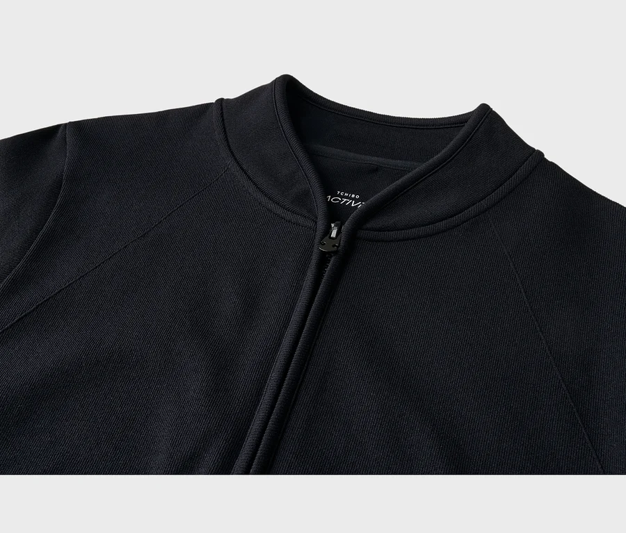 Nahaufnahme einer schwarzen Jacke mit Rippstrickkragen und Reißverschluss, der leicht geöffnet ist. Ein weißes Etikett ist im Nackenbereich sichtbar.