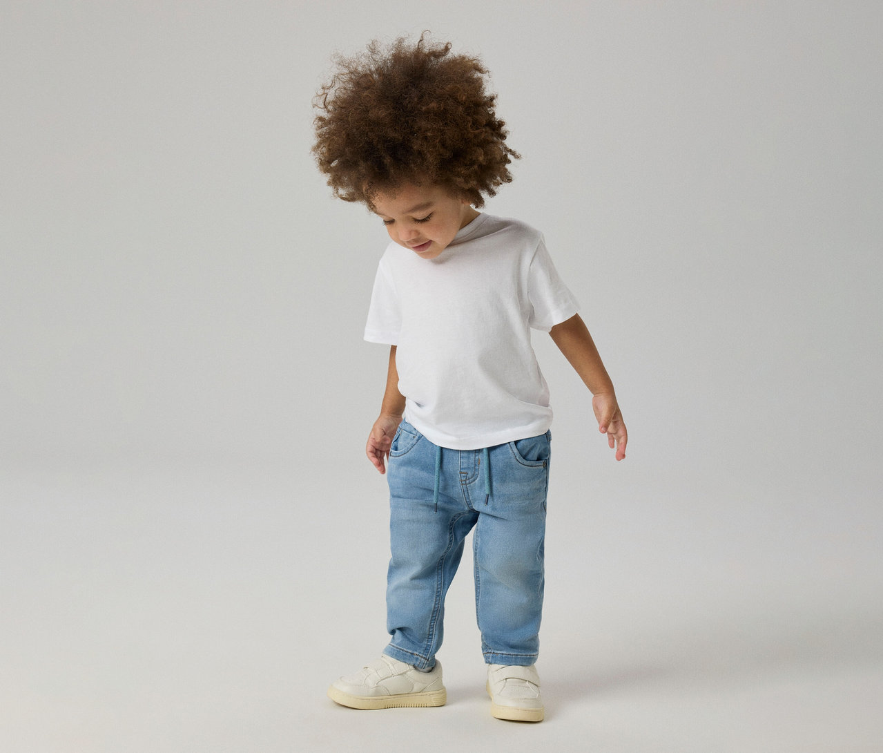 Kleines Kind mit lockigem Haar trägt ein weißes T-Shirt, hellblaue NAME IT Slim Sweat Jeans und helle Sneaker und blickt nach unten.