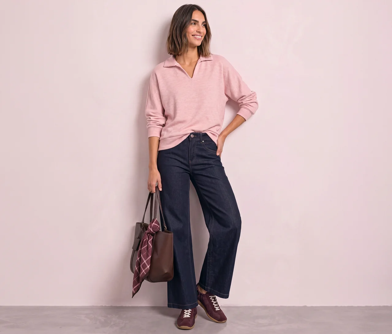 Eine lächelnde Frau mit kurzen braunen Haaren steht, blickt nach rechts und trägt ein hellrosa Street One Poloshirt, dunkelblaue weite Jeans und bordeauxrote Sneaker. Sie hält eine braune Tragetasche mit einem karierten Schal.