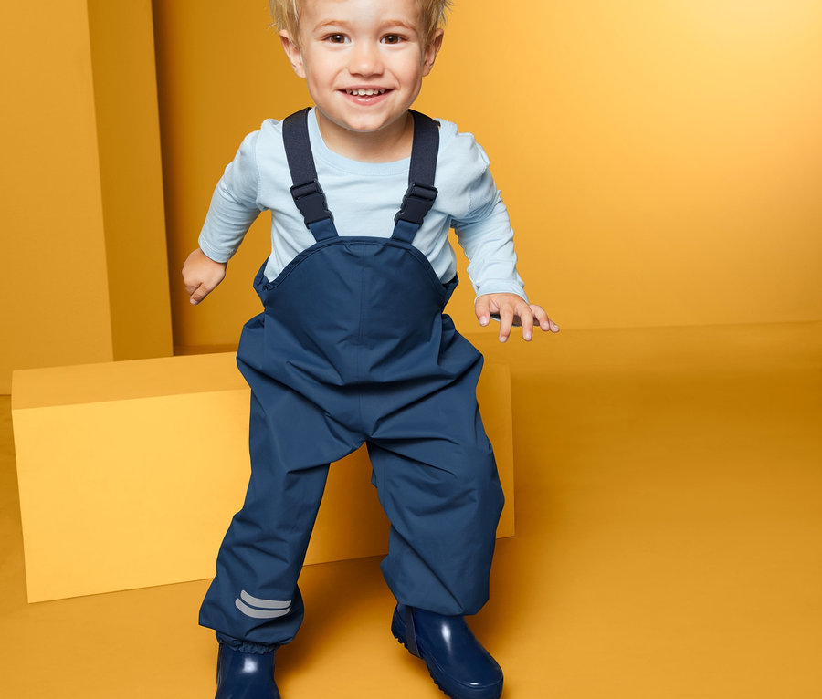 Lächelndes Kleinkind steht vor gelbem Hintergrund und trägt eine blaue Regenhose.