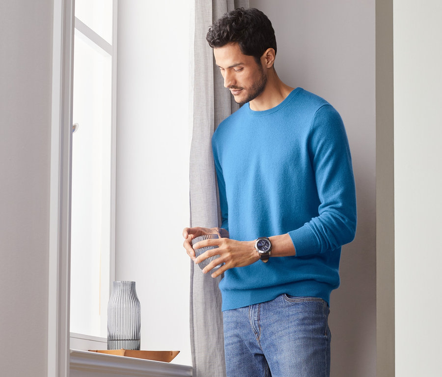 Mann hält ein Glas und schaut aus dem Fenster. Er trägt einen blauen Cashmere-Pullover mit Rundhalsausschnitt.