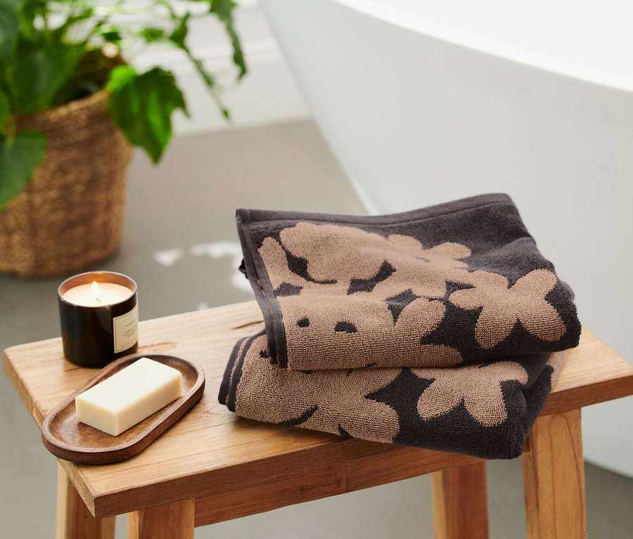 Zwei schwarze Jacquard-Handtücher mit Blumenmuster liegen gefaltet auf einem Holzhocker neben einer Kerze und Seife.
