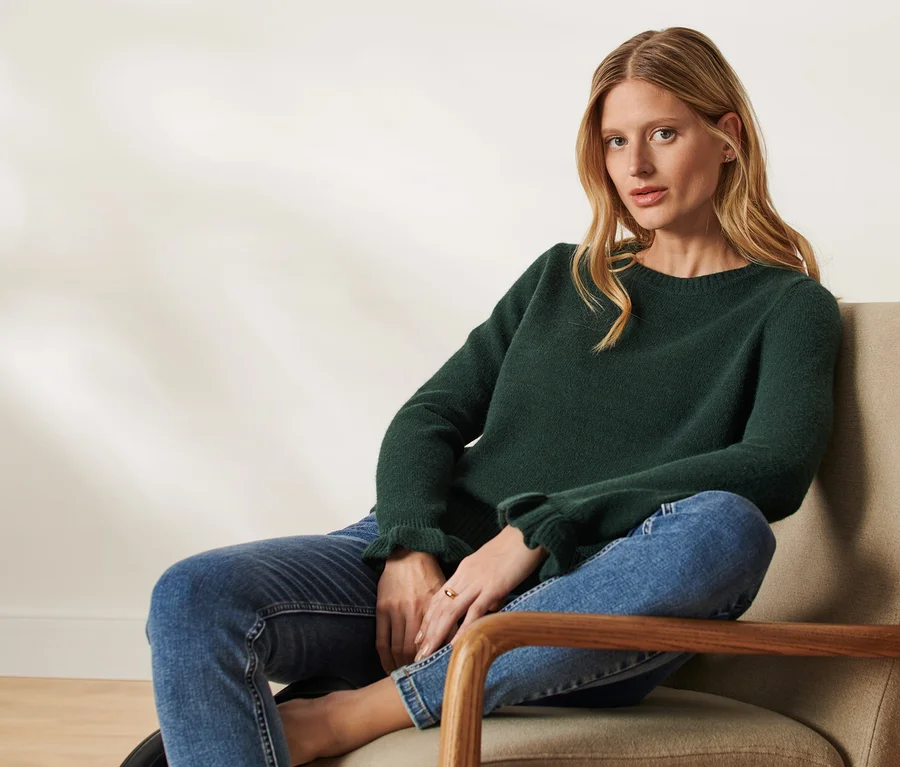 Blonde Frau sitzt im Sessel und trägt einen dunkelgrünen Strickpullover mit Rüschendetail und Jeans.