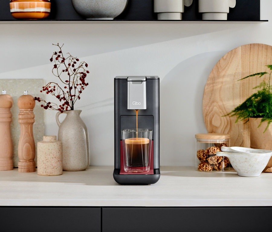 Eine Qbo ICONIC Dark Chrome Kaffeemaschine füllt eine Tasse mit Kaffee. Im Hintergrund Dekorationen.