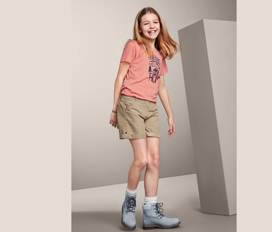 Ein lächelndes Mädchen trägt beige 2-in-1-Funktionsshorts, ein gestreiftes T-Shirt und blaue Stiefel.