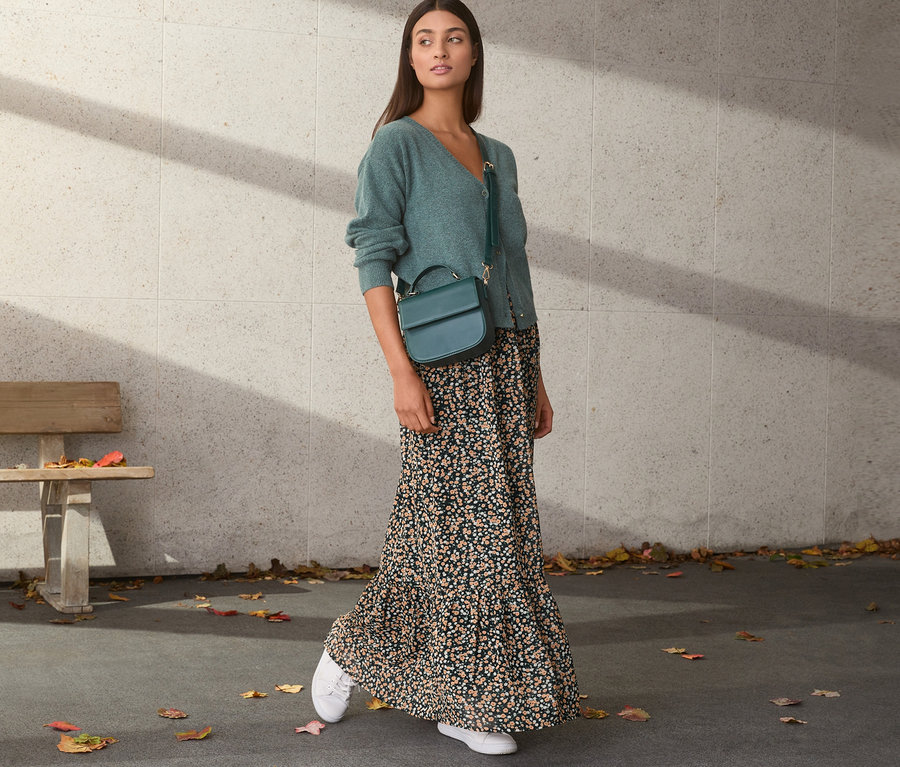 Frau trägt grünen Cardigan und Chiffon-Rock mit Blumenmuster. Sie hat eine grüne Handtasche und weiße Turnschuhe.