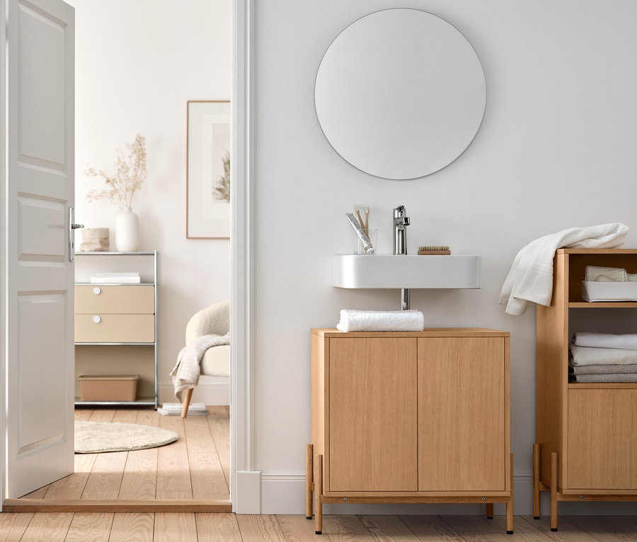 Im Badezimmer stehen ein Waschbeckenunterschrank und ein Badschrank mit Holzfurnier, sowie ein Bouclé-Sessel mit Holzbeinen und ein runder Spiegel »Ameca«.