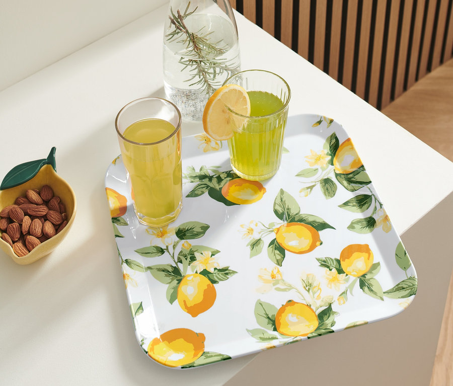 Auf einem weißen Tisch steht ein Tablett mit Zitronenmuster, auf dem sich zwei Gläser mit Getränken und eine Karaffe mit Wasser und Rosmarin befinden. Neben dem Tablett steht eine Schale mit Mandeln.