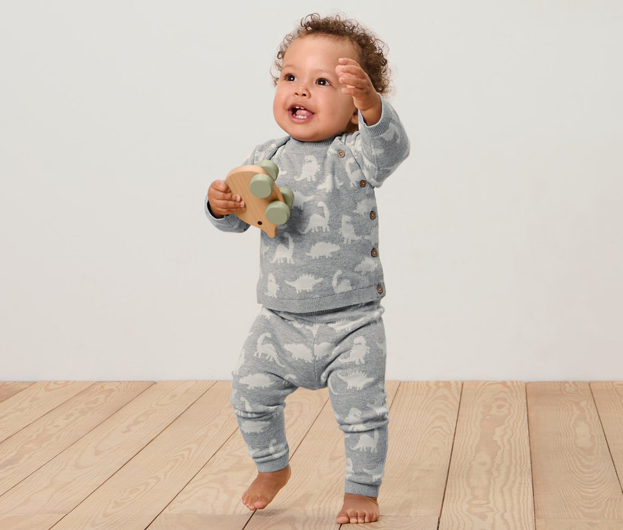 Ein Baby steht auf einem Holzboden und hält ein Holzspielzeug. Es trägt eine Baby-Strickkombi.