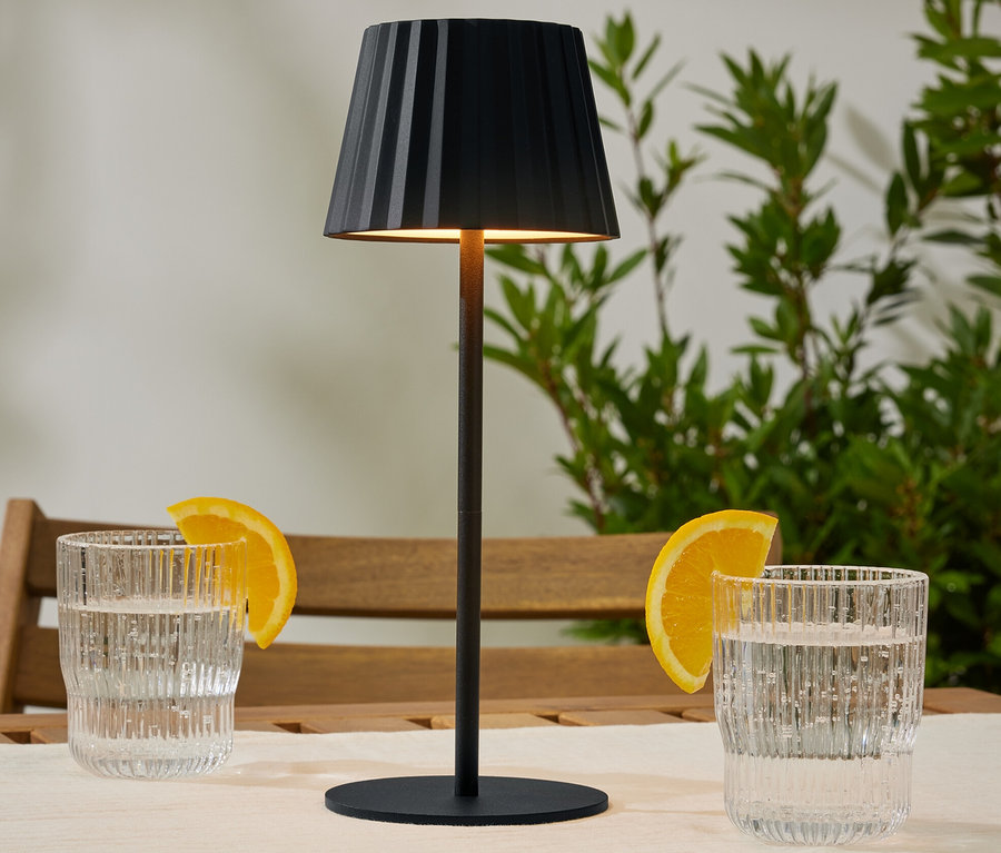 Eine kleine LED-Tischleuchte steht auf einem Holztisch zwischen zwei Gläsern Wasser mit Orangenscheiben.