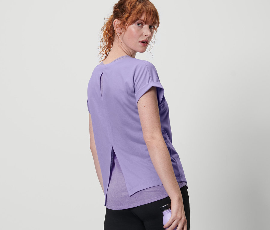 Rothaarige Frau blickt über die Schulter und trägt ein lila 2-in-1-Kurzarm-Sportshirt und schwarze Hose und hält eine lila Wasserflasche.