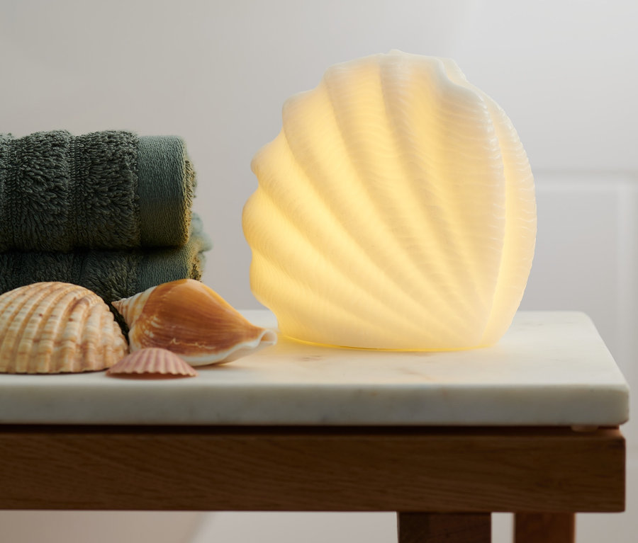Zwei LED-Dekomuscheln stehen auf einem Marmortisch neben grünen Handtüchern und einigen Meeresmuscheln.