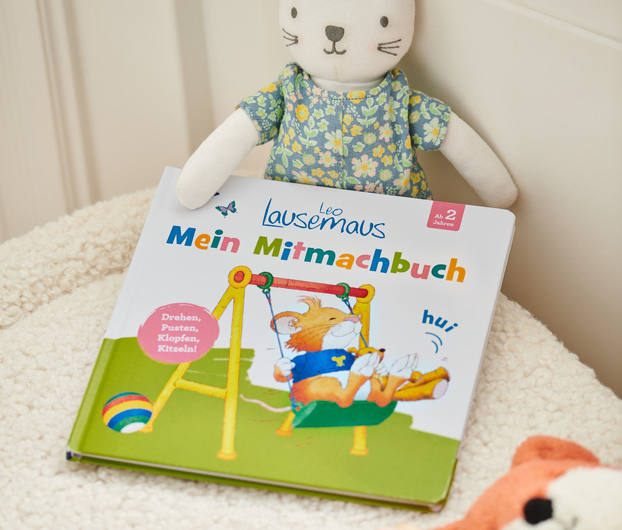 Buch »Leo Lausemaus – mein Mitmachbuch« mit einem Katzenkuscheltier und anderem Spielzeug auf einer zotteligen Oberfläche.