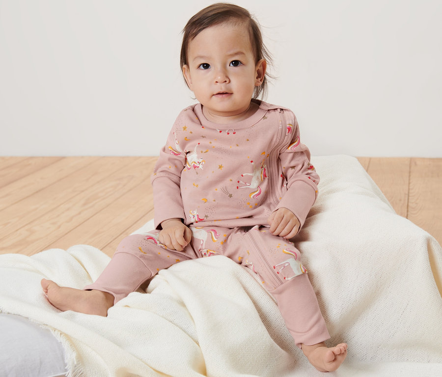 Ein Baby sitzt auf einem Kissen und trägt einen rosa Einhorn-Pyjama.