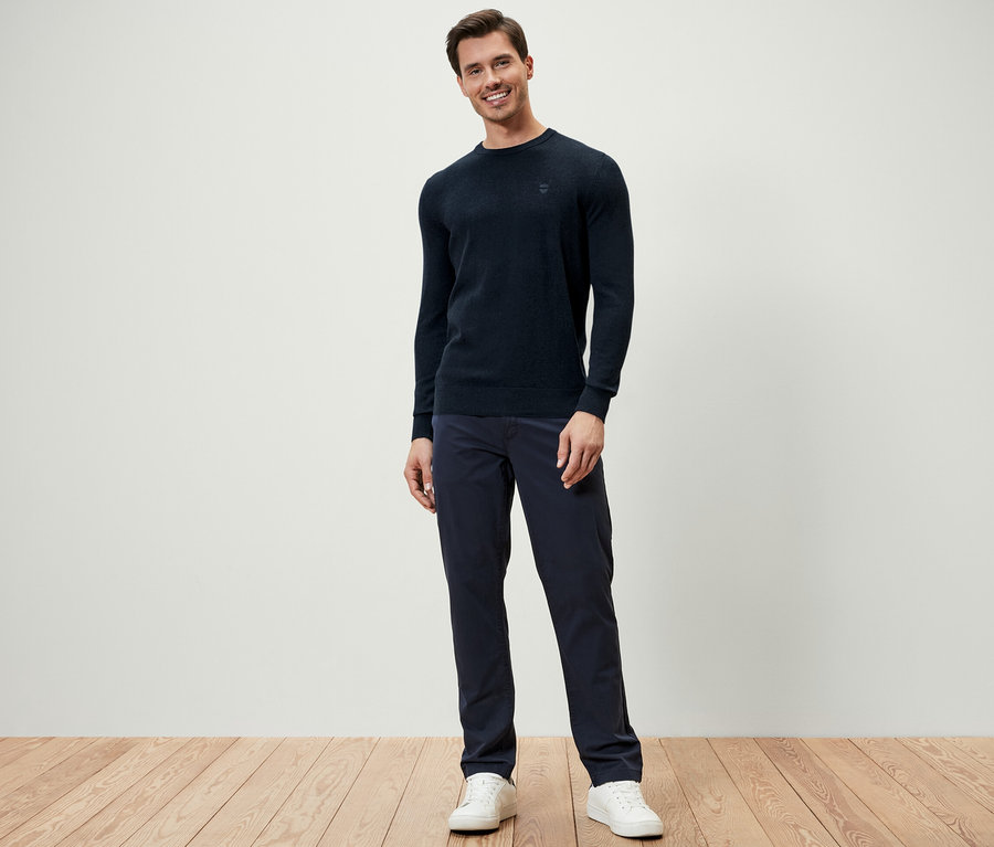 Ein Mann steht und trägt einen dunkelblauen Pullover mit Rundhalsausschnitt, dunkelblaue Hosen und weiße Turnschuhe.