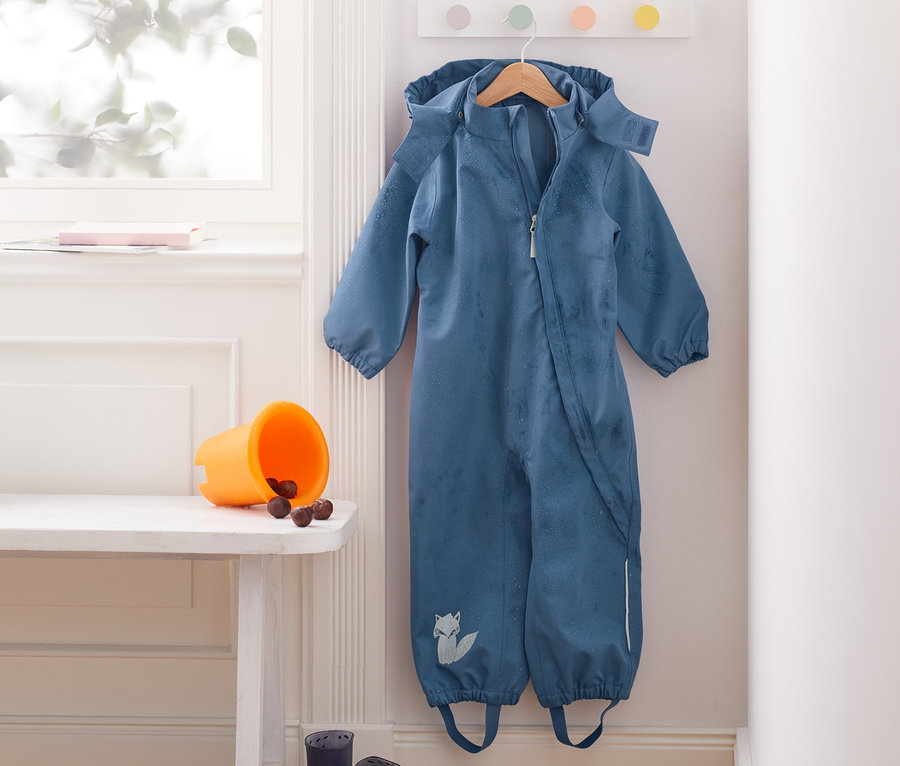 Blauer Regen-Einteiler hängt an einem Kleiderbügel an der Wand. Auf einer Bank stehen ein orangefarbener Eimer mit Kastanien und blaue Gummistiefel. In der Ecke steht eine gelbe Gießkanne.