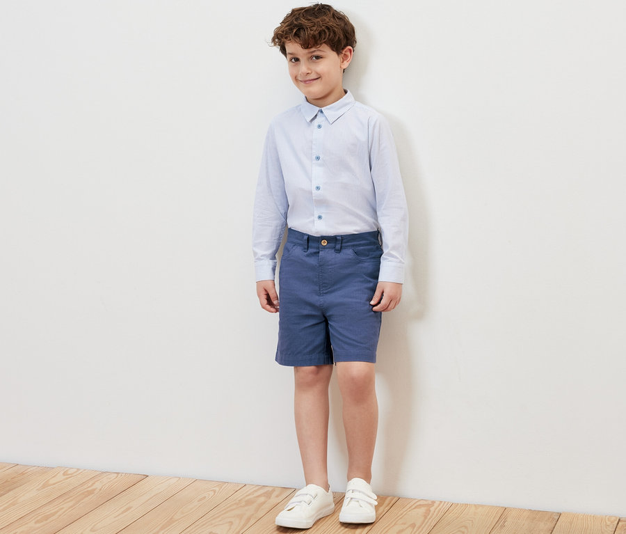 Ein Junge steht und trägt blaue Kinder-Shorts, ein Hemd und Turnschuhe.