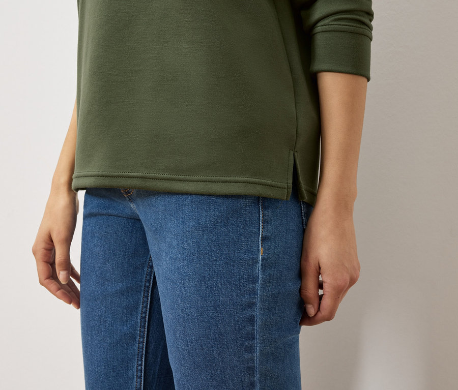 Oberkörper einer Frau in olivgrünem Sweatshirt und blauen Jeans.