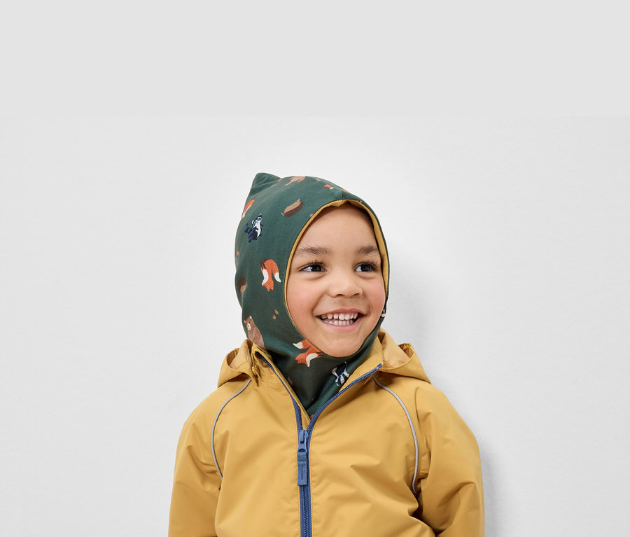 Lächelnder Junge trägt eine grüne Sturmhaube mit Tiermuster und eine gelbe Jacke.