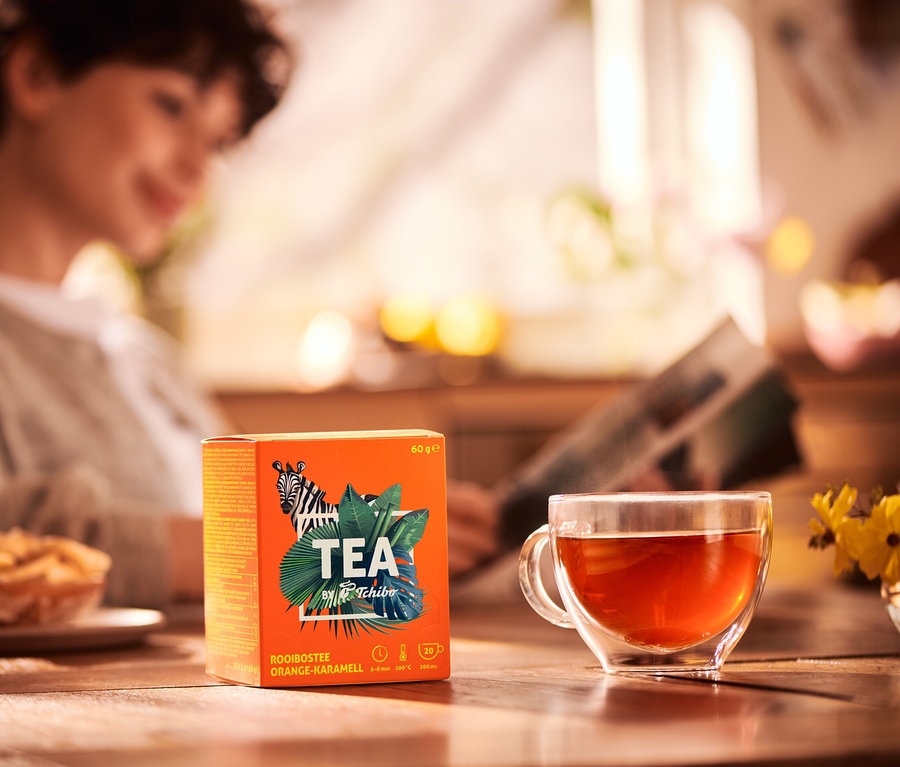 Frau liest Zeitung am Tisch mit Teetasse und Packung TEA by Tchibo Rooibos Orange-Karamell.