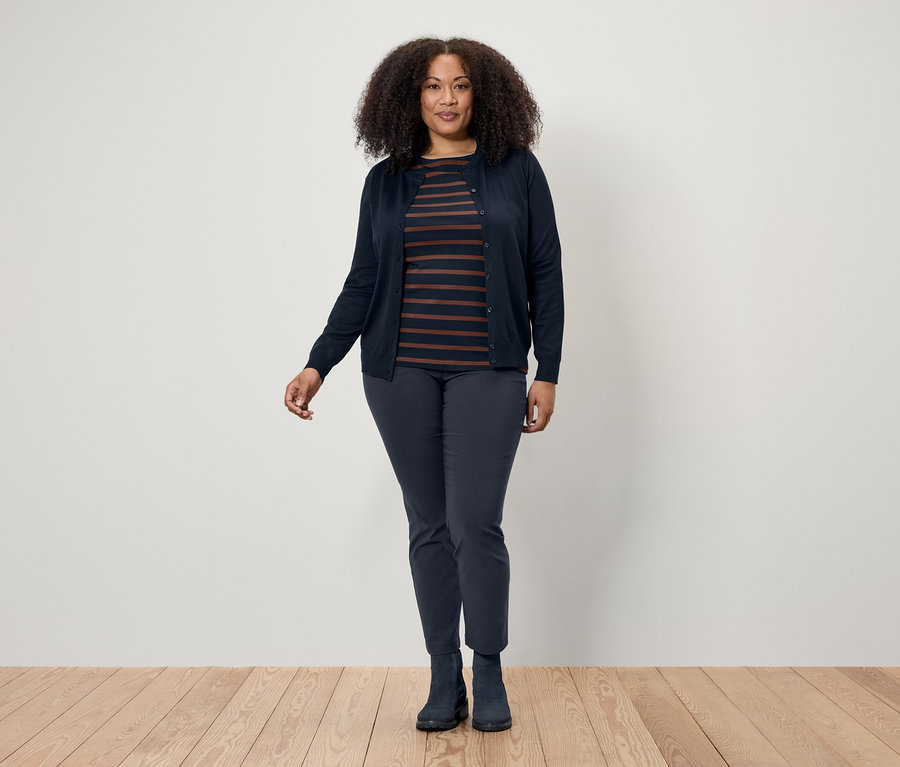 Frau mit dunklem lockigem Haar posiert auf einem Holzboden und trägt eine dunkelblaue Strickjacke, ein gestreiftes T-Shirt und dunkelblaue Hosen und Stiefel.