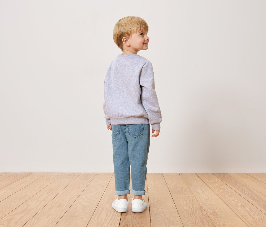 Kleiner Junge mit blonden Haaren, von hinten zu sehen, trägt einen grauen Pullover, blaue Jeans und weiße Schuhe.