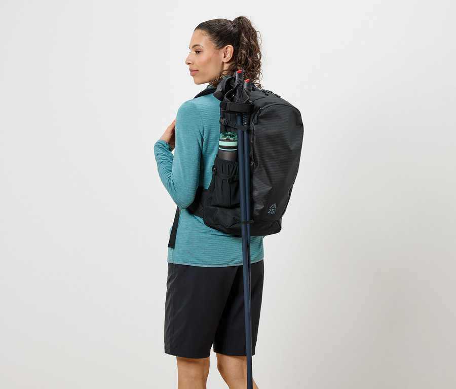 Frau von hinten mit schwarzem Rucksack, Trinkflasche und Wanderstöcken, gekleidet in einem türkisfarbenen Oberteil und schwarzen Shorts.