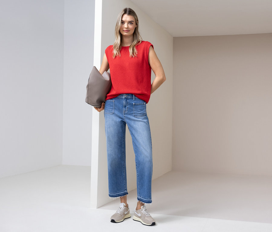 Frau in rotem Top, Jeans und Turnschuhen steht mit einer grauen Tasche in einem hellen Raum.