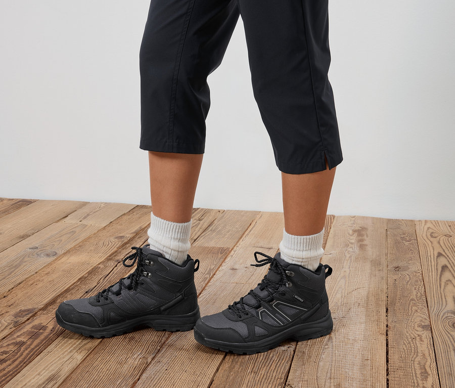 Eine Person trägt schwarze Wanderstiefel und kurze Hosen auf einem Holzboden.