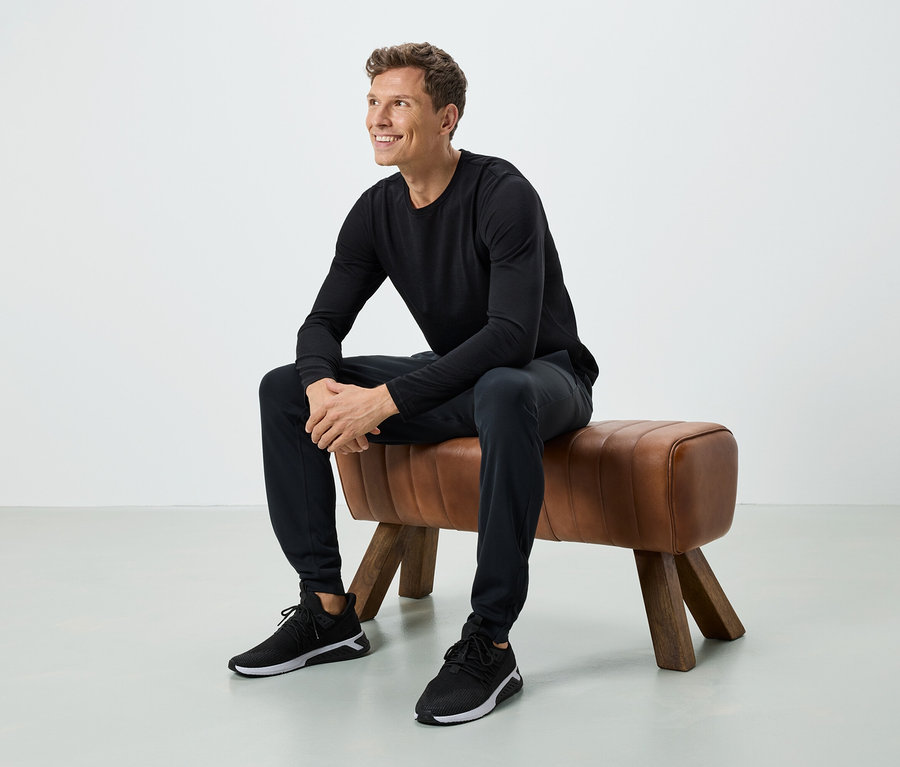 Ein Mann sitzt auf einer braunen Bank. Er trägt ein schwarzes Langarmshirt, eine schwarze Hose und schwarze Turnschuhe.