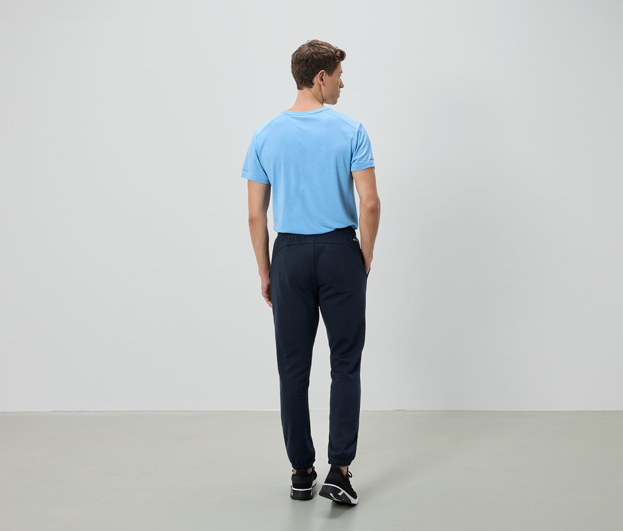 Ein Mann steht mit dem Rücken zur Kamera, er trägt ein hellblaues T-Shirt und eine dunkelblaue Hose.