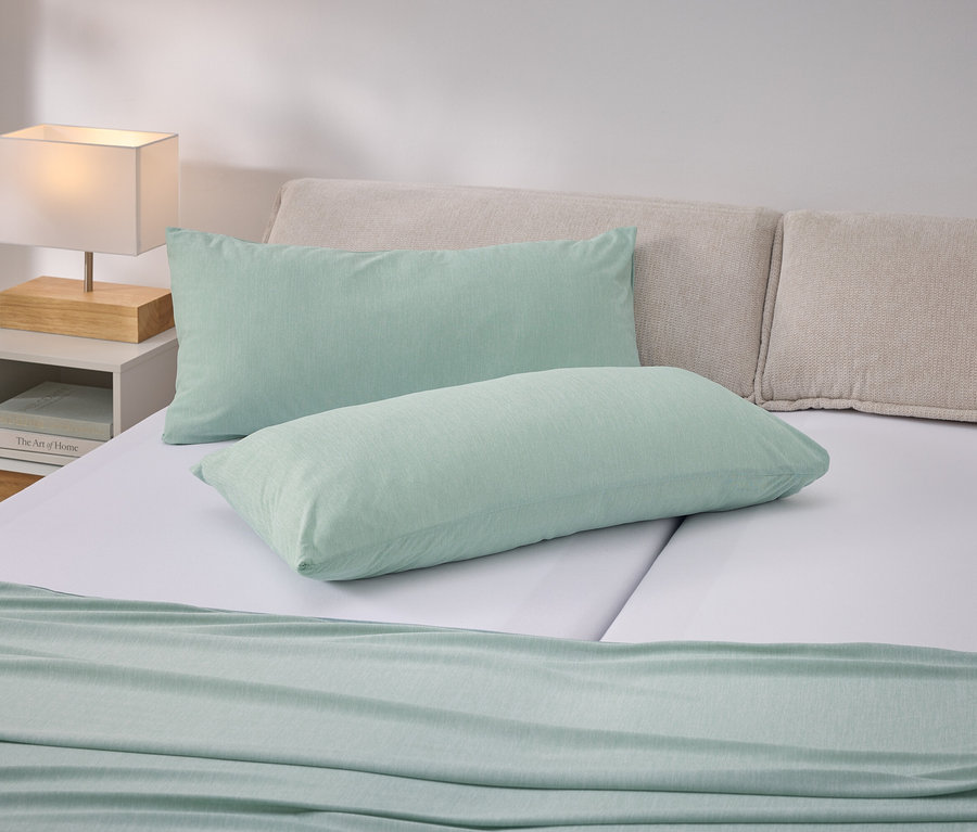 Bett mit hellgrünen Kissen und Tagesdecke auf weißer Bettwäsche. Nachttisch mit Lampe links.