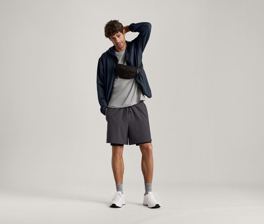 Mann posiert mit der Hand am Kopf, trägt ein hellgrau meliertes Sportshirt, eine dunkle Jacke, Shorts und eine Bauchtasche.