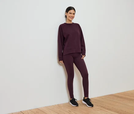 Frau in weinrotem Trainingsanzug, bestehend aus Leggings und Pullover, lehnt an einer weißen Wand.