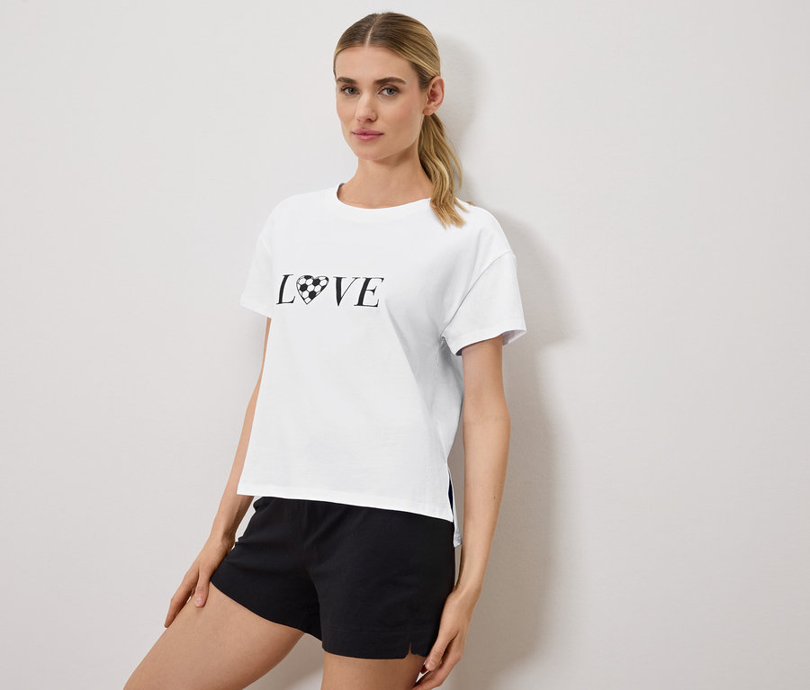 Frau trägt ein weißes T-Shirt mit dem Aufdruck "LOVE", wobei das O durch einen Fußball in Herzform ersetzt wurde, und schwarze Shorts.