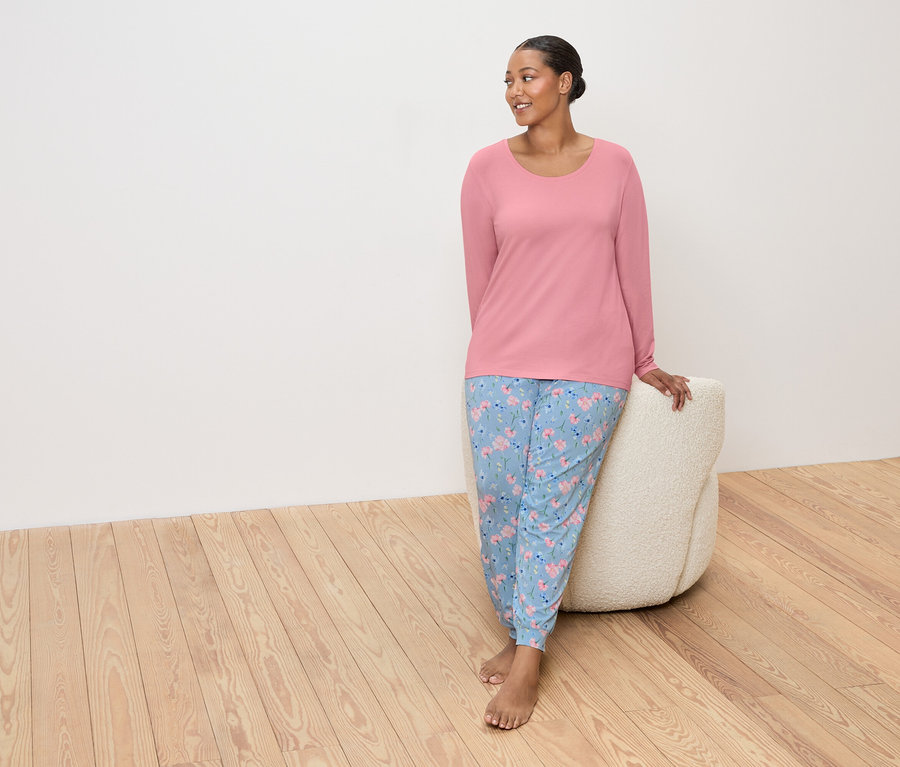 Frau mit rosa Oberteil und blauen, geblümten Hosen, die sich an einen beigen Pouf lehnt.