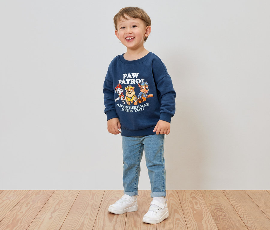 Ein lächelnder Junge trägt ein blaues Paw Patrol-Sweatshirt, Jeans und weiße Turnschuhe.