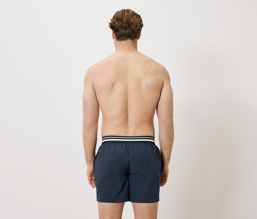Rückansicht eines Mannes mit freiem Oberkörper, der dunkelblaue Badeshorts mit weißem Streifen trägt.