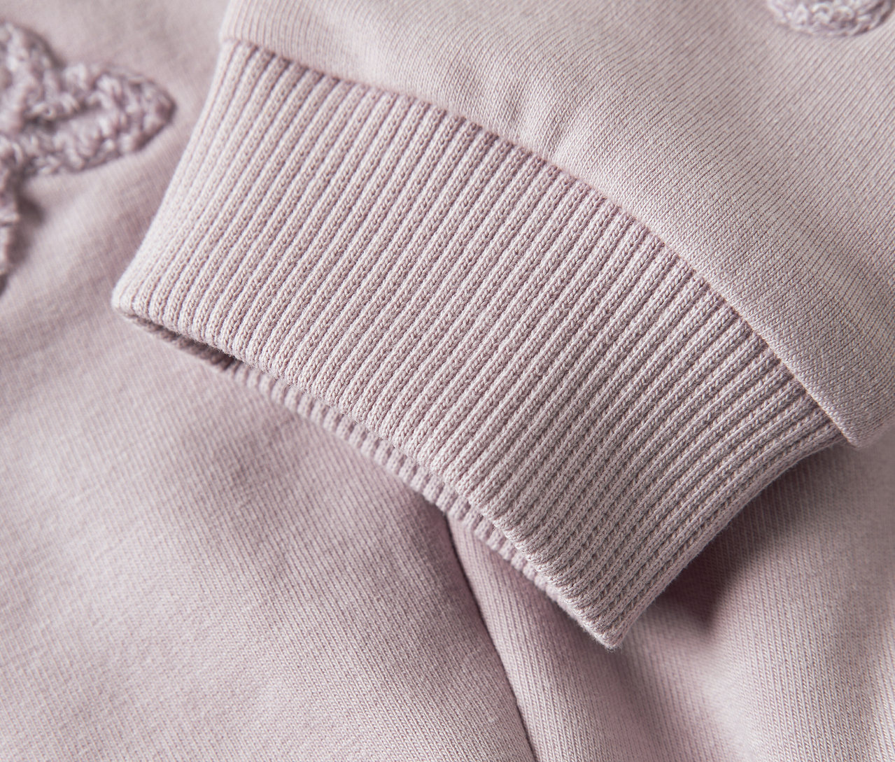 Nahaufnahme vom Ärmel eines lila Sweatshirts mit Rippbündchen und Blumenapplikation.